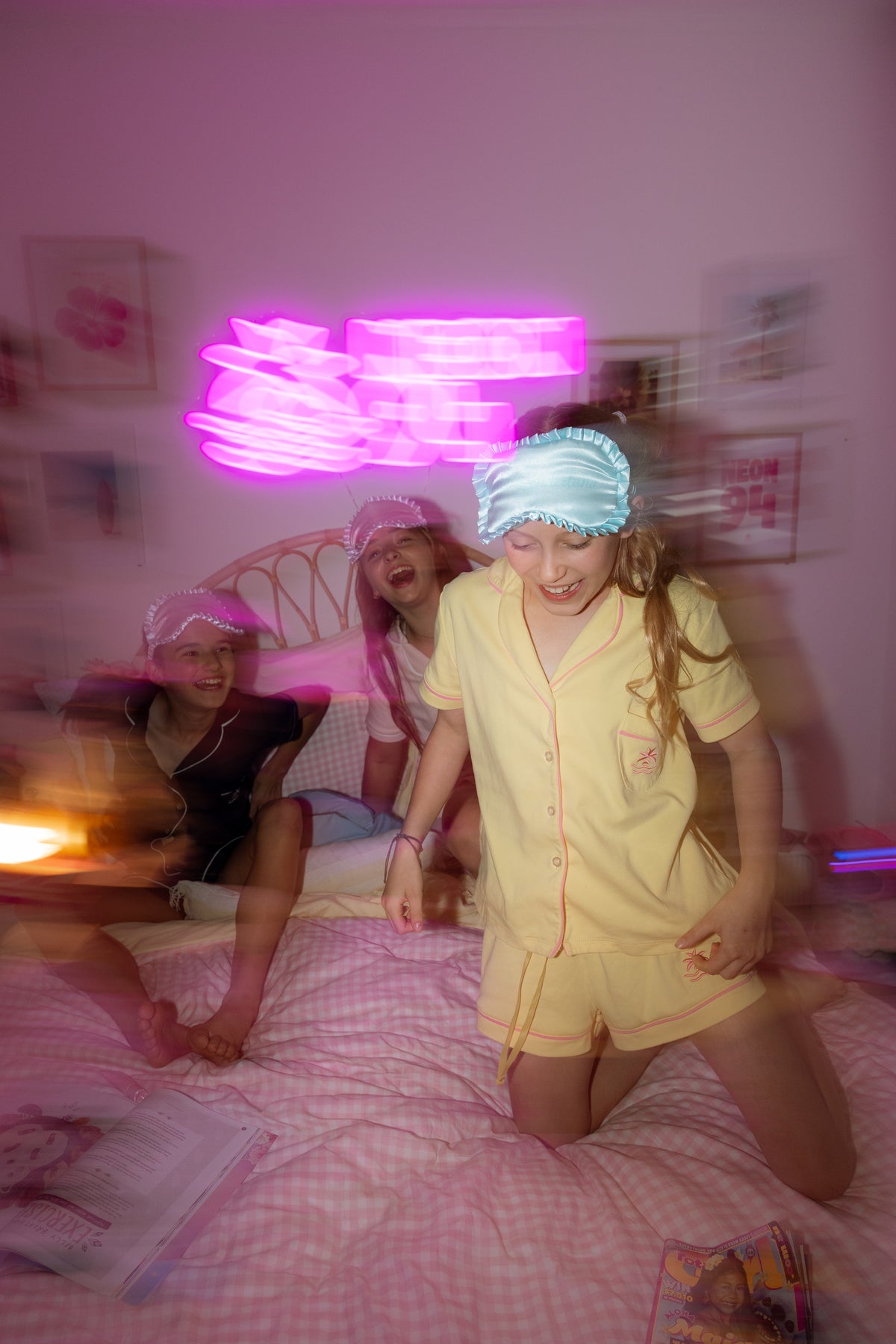 Girl in the Neon94 Popcorn butter yellow pyjama set dancing on a bed under a pink neon sign